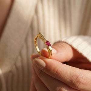 May include: A gold ring featuring a rectangular, red gemstone. The ring has a modern design with a minimalist band. The red gemstone is set horizontally, adding a pop of colour to the gold band. The ring is held up by a person.