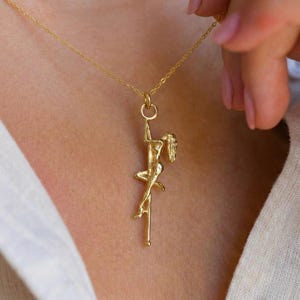 Può includere: Una collana con pendente dorato raffigurante una figura su un palo. Il pendente è sospeso a una catena sottile. La figura è in una posa dinamica, con i capelli fluenti. La collana è presentata su uno sfondo chiaro. Il pendente è color oro.
