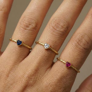 Op de afbeelding: Drie gouden ringen, elk met een hartvormige edelsteen. Eén ring heeft een blauwe edelsteen, een andere een heldere edelsteen en de derde een rode edelsteen. De ringen worden getoond op de vingers van een persoon.