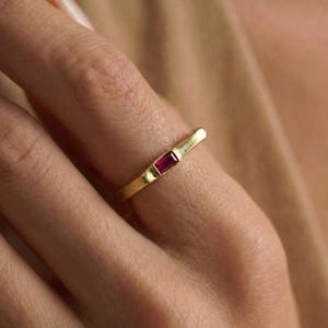 May include: A gold ring featuring a rectangular, deep red gemstone. The ring is on a finger, highlighting the minimalist design and the vibrant colour of the stone. The band has a textured finish.