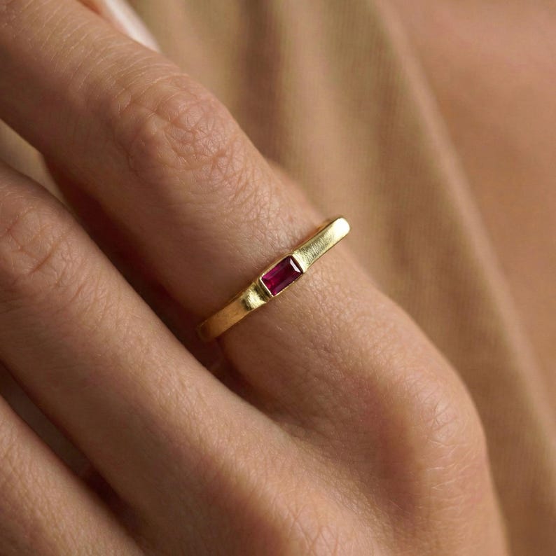 May include: A gold ring featuring a rectangular, deep red gemstone. The ring is on a finger, highlighting the minimalist design and the vibrant colour of the stone. The band has a textured finish.