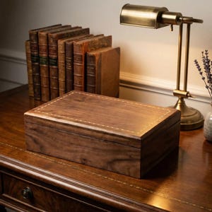 May include: A rectangular wooden box with a decorative stitched border sits on a dark wooden desk. The box is surrounded by antique books, a brass desk lamp, and a small vase with lavender sprigs. The box is a rich brown color.