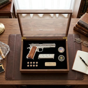 May include: A Colt Model 1911 pistol displayed in a wooden case with a glass top. The case contains the pistol, a magazine, several bullets, and two commemorative coins. The pistol has a silver finish and a wooden grip.