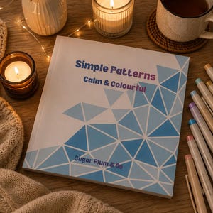 Può includere: Un libro intitolato "Simple Patterns, Calm & Colourful" con un design geometrico blu. Il libro è su una superficie di legno, circondato da candele, una tazza di tè e penne colorate. La copertina del libro include anche il testo "Sugar Plum & Co."
