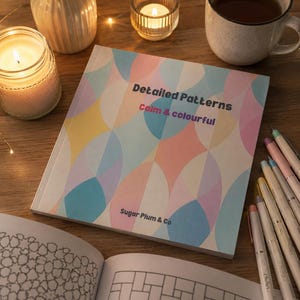 Op de afbeelding: Een kleurboek getiteld "Detailed Patterns Calm & Colourful" met een geometrisch ontwerp op de omslag. Het boek is open op een pagina met een geometrisch patroon. Kleurpotloden, kaarsen en een kopje thee staan ook op het houten oppervlak.