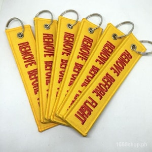 May include: A collection of yellow fabric keychains with the embroidered text "REMOVE BEFORE FLIGHT" in red. Each keychain has a silver metal ring attached for easy use. The keychains are rectangular and have a stitched border.