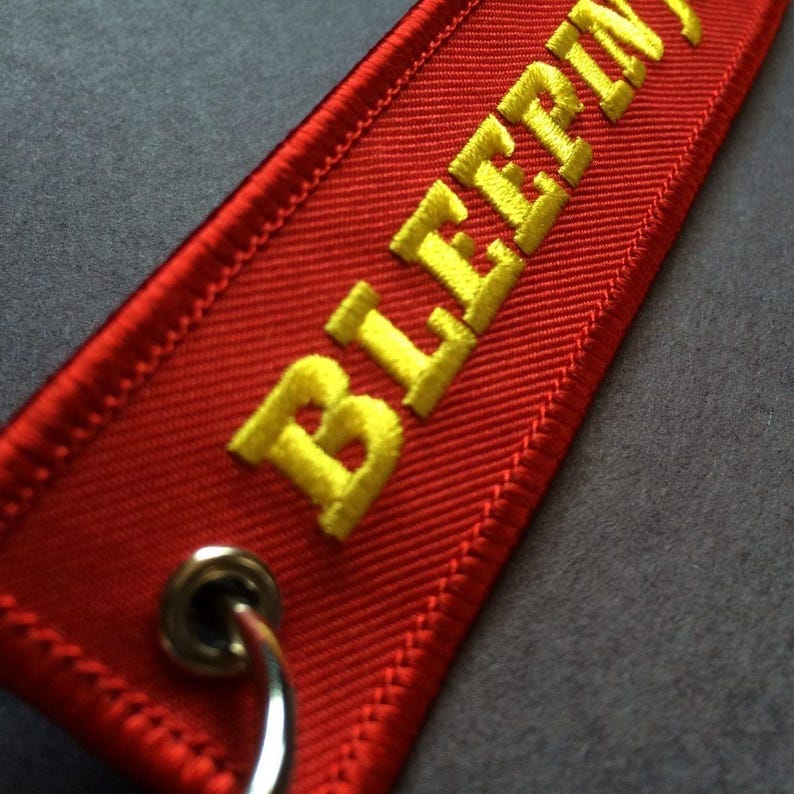 May include: Red fabric keychain tag with yellow embroidered text that reads "BLEEPIN". The tag has a metal grommet and a silver key ring attached. The edges are neatly stitched.