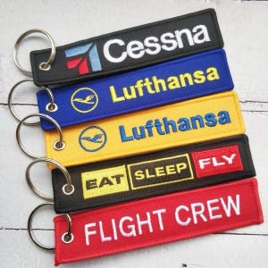 May include: Embroidered keychains in black, blue, yellow, and red. The keychains feature the text "Cessna", "Lufthansa", "EAT SLEEP FLY", and "FLIGHT CREW" in white or yellow. Each keychain has a silver metal ring.