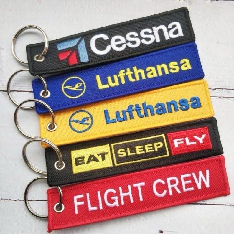 May include: Embroidered keychains in black, blue, yellow, and red. The keychains feature the text "Cessna", "Lufthansa", "EAT SLEEP FLY", and "FLIGHT CREW" in white or yellow. Each keychain has a silver metal ring.