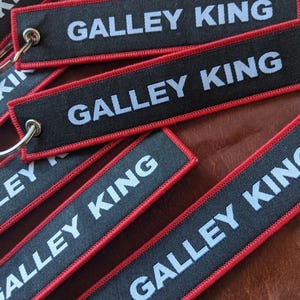 May include: Black rectangular keychains with the text "GALLEY KING" in white, outlined with red stitching. Each keychain has a silver metal ring attached for keys. The keychains are scattered on a brown surface.