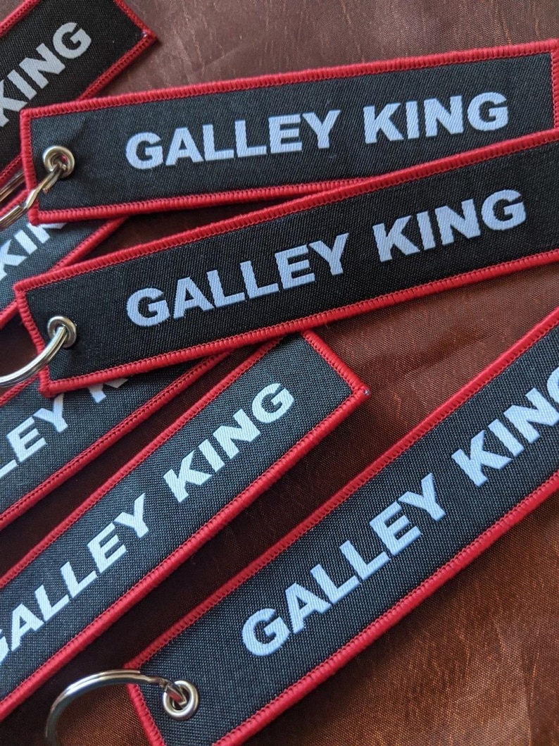 May include: Black rectangular keychains with the text "GALLEY KING" in white, outlined with red stitching. Each keychain has a silver metal ring attached for keys. The keychains are scattered on a brown surface.
