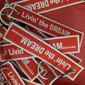 May include: A collection of red keychains with white embroidered text that reads "Livin' the DREAM." Each keychain has a white border and a silver metal ring. The keychains are scattered on a red surface.