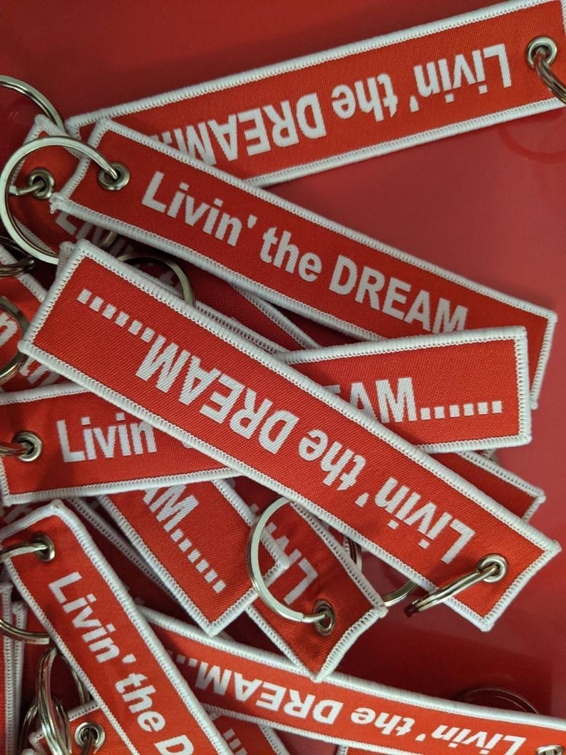 May include: A collection of red keychains with white embroidered text that reads "Livin' the DREAM." Each keychain has a white border and a silver metal ring. The keychains are scattered on a red surface.