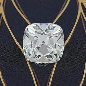 May include: A square, clear cushion-cut diamond with many facets, set against a dark blue background with a gold leaf pattern. The diamond is highly reflective, showcasing its brilliance.