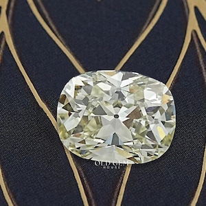 May include: A cushion-cut diamond with a light yellow hue. The gemstone is set against a dark blue background with a gold leaf pattern. The diamond's facets reflect light, creating a sparkling effect.