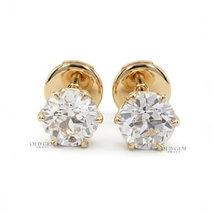 May include: A pair of stud earrings featuring round, faceted clear stones set in yellow gold prongs. Each earring has a post back mechanism. The text "OLD GEM HOUSE" is printed in small black letters near the base of each earring.