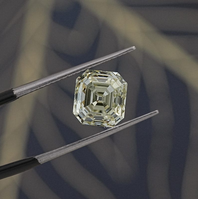 May include: A square-cut, light yellow diamond held by tweezers. The gemstone has a complex, multi-faceted design, reflecting light. The background is a blurred mix of gray and blue tones, providing contrast to the diamond's brilliance.