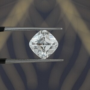May include: A brilliant, cushion-cut diamond held by tweezers. The colorless gemstone reflects light, showcasing its facets. The diamond's shape is square with rounded corners, a classic choice for jewelry.