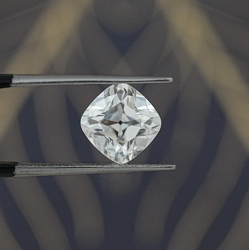 May include: A brilliant, cushion-cut diamond held by tweezers. The colorless gemstone reflects light, showcasing its facets. The diamond's shape is square with rounded corners, a classic choice for jewelry.