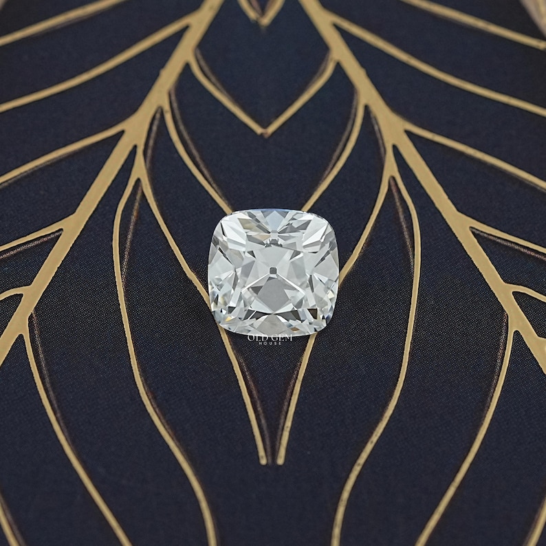May include: A square, clear cushion-cut diamond with many facets, set against a dark blue background with a gold leaf pattern. The diamond is highly reflective, showcasing its brilliance.