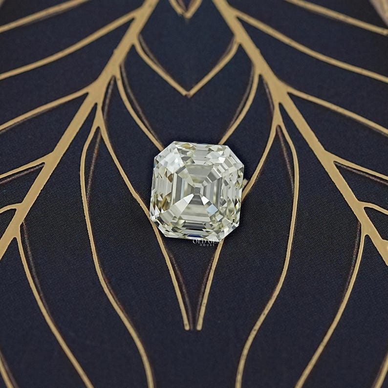 May include: A large, square-cut diamond with multiple facets, resting on a dark blue fabric with a gold leaf pattern. The diamond reflects light, showcasing its clarity and brilliance. The overall composition suggests luxury and elegance.
