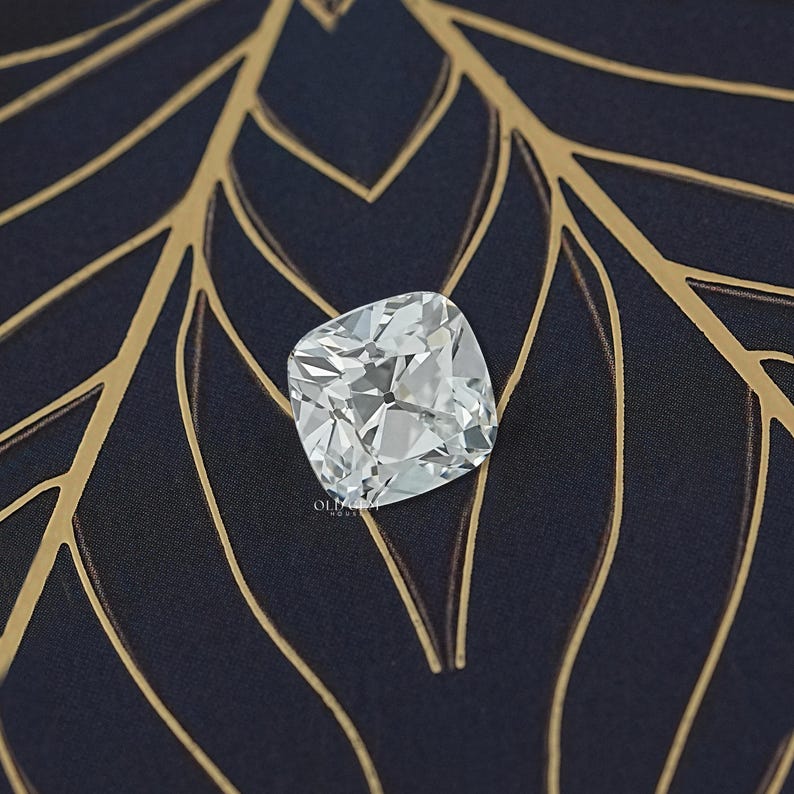 May include: A square-cut, clear diamond with multiple facets, reflecting light. The diamond is set against a dark blue fabric with a gold leaf pattern. The image is well-lit, highlighting the diamond's brilliance.