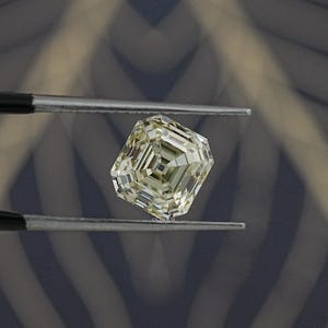 May include: A faceted, light yellow diamond held by tweezers. The diamond has an emerald cut with multiple facets, reflecting light. The background is a blurred pattern of gray and blue.