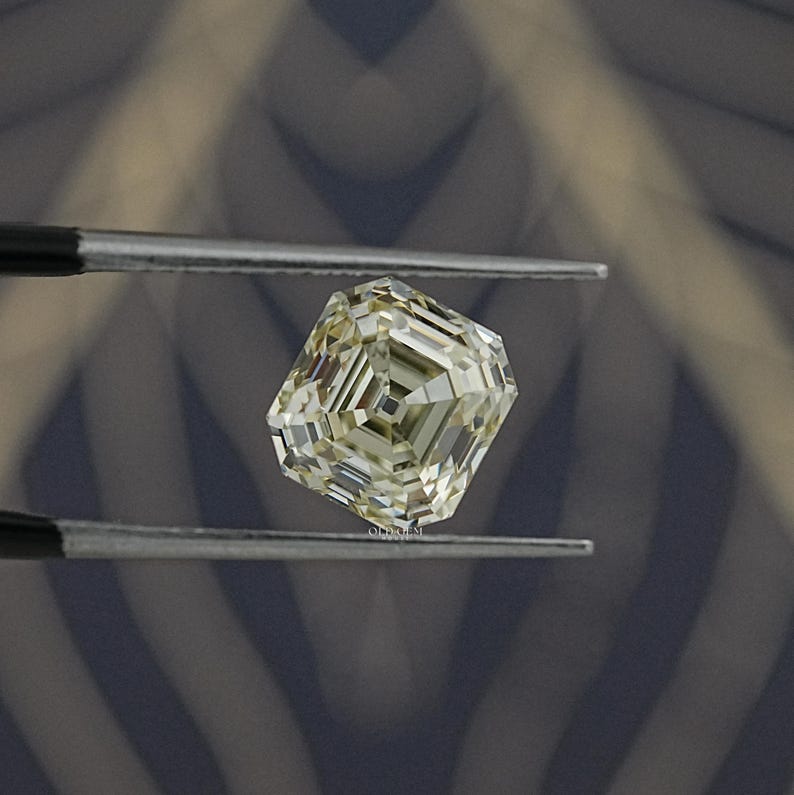 May include: A faceted, light yellow diamond held by tweezers. The diamond has an emerald cut with multiple facets, reflecting light. The background is a blurred pattern of gray and blue.