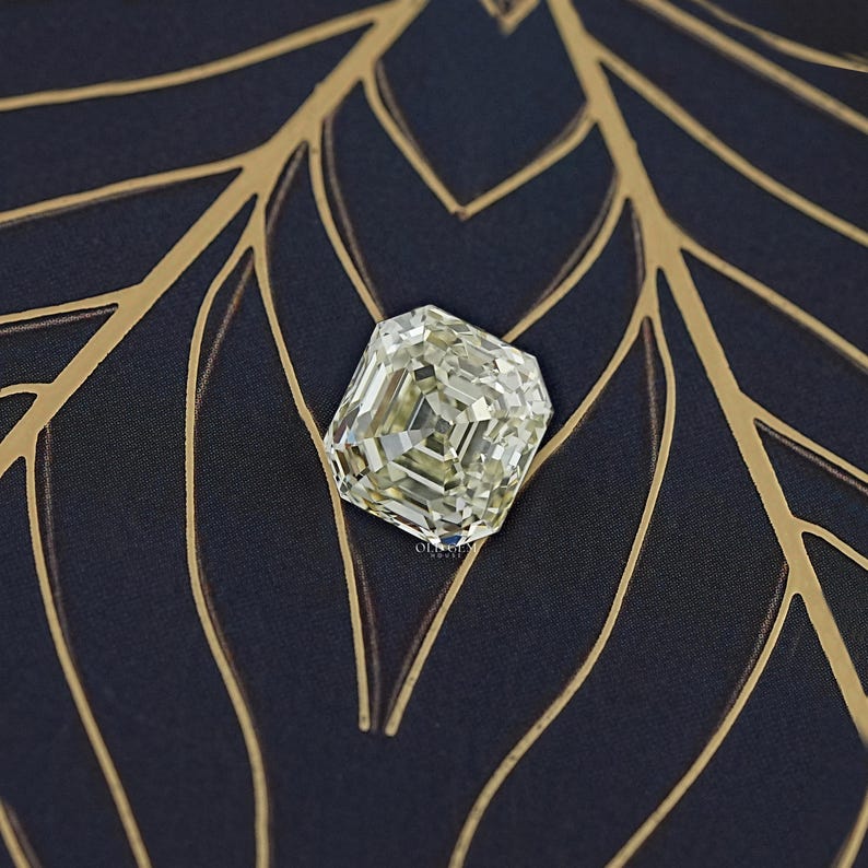 May include: A large, rectangular-cut diamond with a pale yellow hue. The gemstone is set against a dark blue fabric with a gold leaf pattern. The diamond's facets reflect light, creating a sparkling effect.