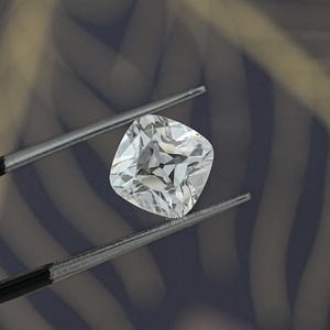 May include: A square-cut, clear gemstone held by tweezers. The gem has multiple facets that reflect light, creating a sparkling effect. The background is a blurred mix of dark and light tones.