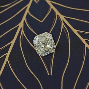 May include: A large, square-cut diamond with a pale yellow hue. The gemstone is set against a dark blue background with gold leaf-like patterns. The diamond's facets reflect light, creating a sparkling effect.