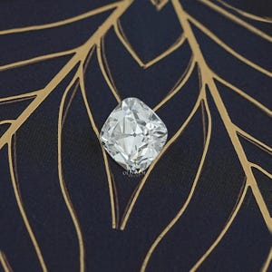 May include: A large, square-cut, clear gemstone with many facets, reflecting light. The gem is set against a dark blue fabric with a gold leaf pattern. The image is well-lit, highlighting the brilliance of the stone.