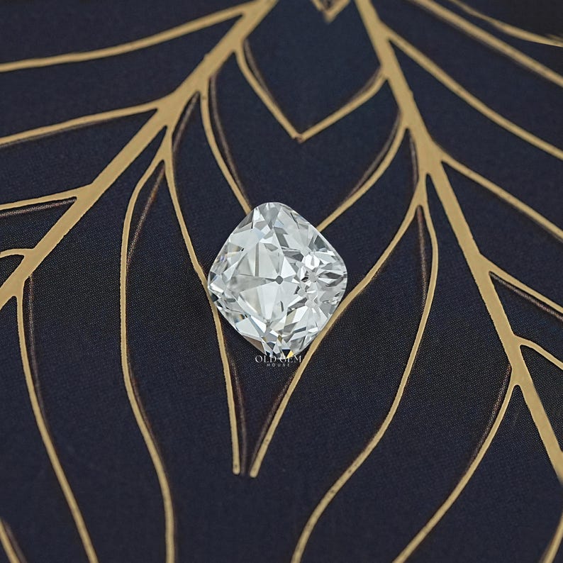 May include: A large, square-cut, clear gemstone with many facets, reflecting light. The gem is set against a dark blue fabric with a gold leaf pattern. The image is well-lit, highlighting the brilliance of the stone.