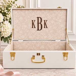 May include: A white rectangular storage box with gold-toned metal latches and a central handle. The interior is lined with a beige fabric featuring a white floral pattern, and the inside lid displays the embroidered monogram letters KBK in brown.