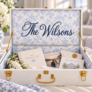 May include: A white wooden keepsake box with gold latches and a handle. The interior is lined with blue and white floral fabric. The lid features the text The Wilsons. Inside are a wedding album, a photograph, and a Save the Date card.