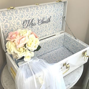May include: A white rectangular storage chest with gold-tone metal latches and hinges. The interior is lined with a light blue and white floral pattern fabric. The text "Mrs Smith" is printed in black script on the inside lid. A bouquet of cream and pink artificial roses with a white tulle veil is placed inside the chest.