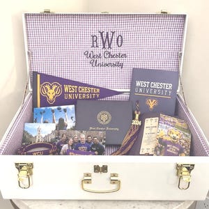 May include: A white keepsake trunk with gold latches and a purple gingham interior lining, filled with West Chester University memorabilia including a pennant, a diploma cover, photographs, a 2024 tassel, and a mascot image with university text.