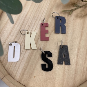Può includere: Una collezione di portachiavi con lettere colorate, tra cui bianco, malva, blu navy, nero e grigio antracite. Ogni lettera è attaccata a un portachiavi argentato. I portachiavi sono disposti su un vassoio di legno chiaro.