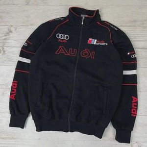 May include: Black zip-up jacket with red and white accents. Features the Audi logo and text in red on the chest and sleeves. Includes "Audi Sports" and "Audi Lounge" text. A sporty, casual jacket.