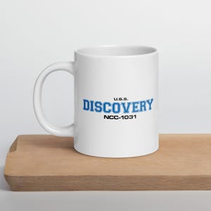 May include: White ceramic mug featuring the text "U.S.S. DISCOVERY NCC-1031" in blue and black. The mug has a large handle and is on a wooden surface. The mug is a Star Trek themed product.
