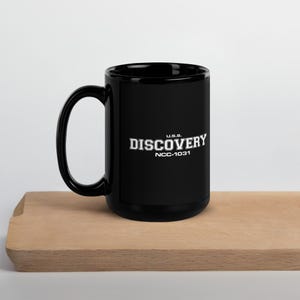 May include: Black ceramic mug featuring the text "U.S.S. DISCOVERY NCC-1031" in white. The mug has a large handle and is on a wooden surface. The mug is a Star Trek themed product.