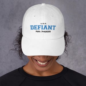 Könnte beinhalten: Weiße Baseballkappe mit dem Text "U.S.S. DEFIANT NX-74205" in blauer Schrift. Die Kappe hat einen gebogenen Schirm und ein klassisches Sechs-Panel-Design. Der Hut ist aus einem weichen Material.
