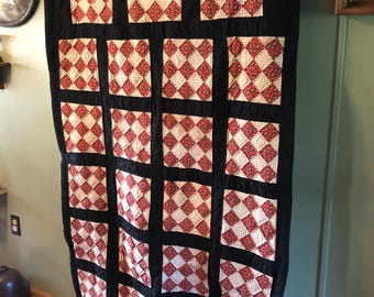 May include: A handmade quilt with a black border and a repeating pattern of red and cream-colored squares. The quilt is hanging on a rod, showcasing its design and craftsmanship. The quilt's dimensions are not visible.