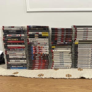以下が含まれることがあります： ベージュ色の表面に積み重ねられたPlayStation 3のビデオゲームケースのコレクション。タイトルには、NBA 2K、NHL 2K、Pro Evolution Soccerなどがあります。ケースは透明なプラスチック製で、さまざまなゲームカバーが特徴です。