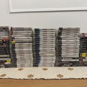 May include: A large collection of PlayStation 3 game cases, mainly from the FIFA Soccer series, arranged in stacks. The cases are white with game titles and logos. Other game titles are also visible.