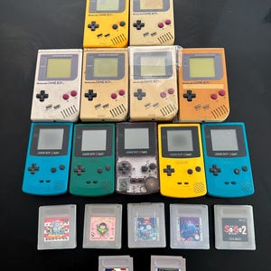 以下が含まれることがあります： 黄色、ベージュ、オレンジ、青、緑など、様々な色のビンテージGame BoyとGame Boy Colorの携帯ゲーム機コレクション。ゲームタイトルとグラフィックが特徴のゲームカートリッジもいくつか見えます。