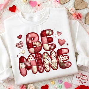 May include: White sweatshirt featuring the phrase "BE MINE" in a patchwork design. The letters are red and pink with various patterns. Hearts and a Valentine's Day card are also visible.