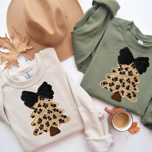 Può includere: Due felpe girocollo, una color crema e una verde oliva, ciascuna con un design ad albero di Natale con stampa leopardata e un fiocco nero. Un cappello beige, una foglia autunnale e una tazza di caffè completano il tema stagionale.