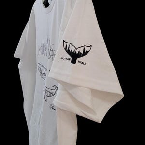 Puede incluir: Camiseta blanca con un dise&ntilde;o gr&aacute;fico negro de una cola de ballena y el texto "Gotham Whale" en la manga. La camiseta tambi&eacute;n tiene un dise&ntilde;o de ballena y horizonte urbano. La camiseta est&aacute; colgada en una percha de pl&aacute;stico transparente.