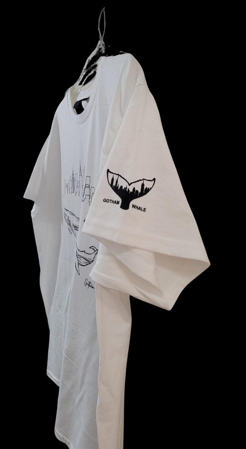 Puede incluir: Camiseta blanca con un dise&ntilde;o gr&aacute;fico negro de una cola de ballena y el texto "Gotham Whale" en la manga. La camiseta tambi&eacute;n tiene un dise&ntilde;o de ballena y horizonte urbano. La camiseta est&aacute; colgada en una percha de pl&aacute;stico transparente.